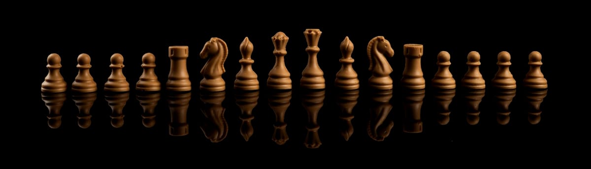 The light chocolate pieces of a Caissa chess set standing in a row on a black, reflective surface: pawns, rooks, knights, bishops, the queen and the king.